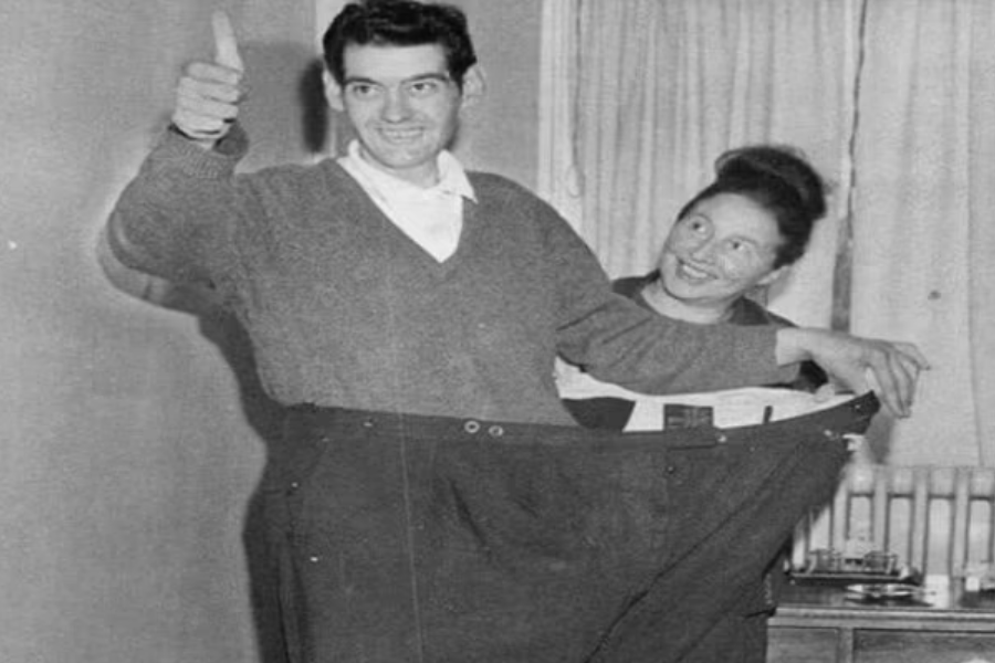 Smiling man giving a thumbs up while standing in dramatically oversized trousers, with a woman beside him holding the waistband.