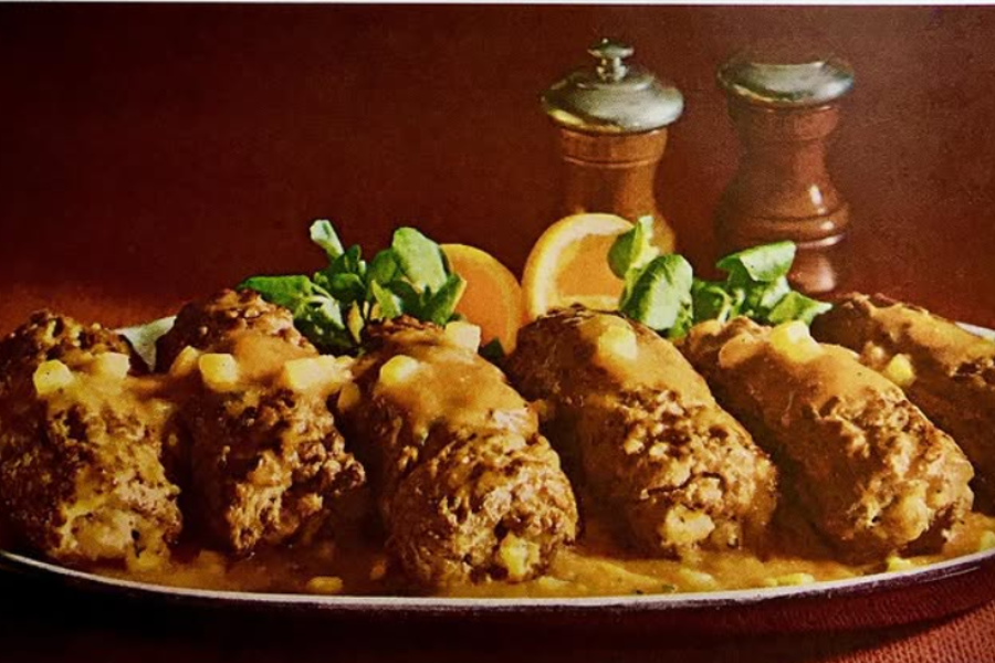 A platter of beef roulades covered in brown gravy, served with leafy greens, orange slices, and garnished with diced vegetables.