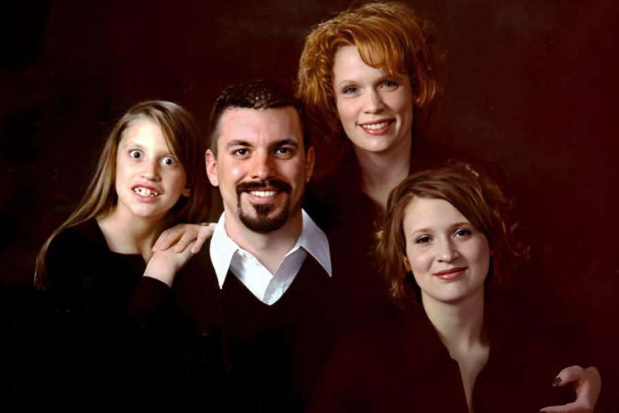 A professionally lit family portrait featuring four people in black outfits, with one young girl on the left making an exaggerated wide-eyed face.