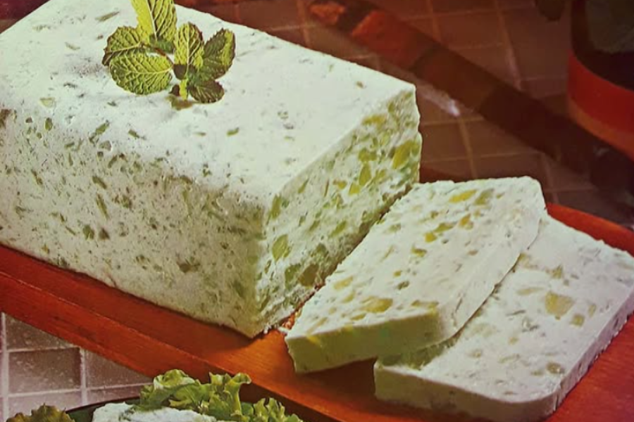 A pale green, loaf-shaped gelatin mold with bits of pineapple and what appears to be celery, garnished with a mint sprig on top and sliced on the side.