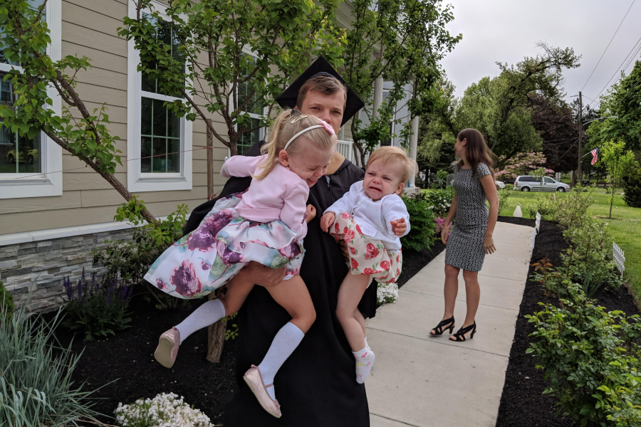 A man in a graduation gown holds two crying toddlers in floral dresses, while a woman walks away in the background, seemingly over the chaos.