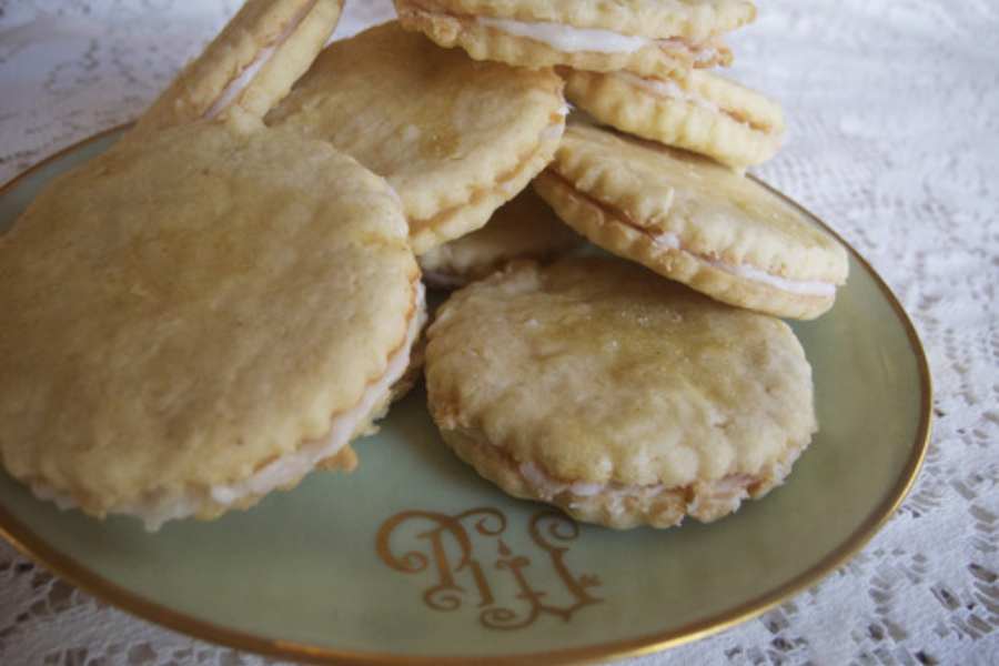 Plate of round, golden-brown coconut sandwich cookies with cream filling, arranged on a vintage dish with ornate lettering.