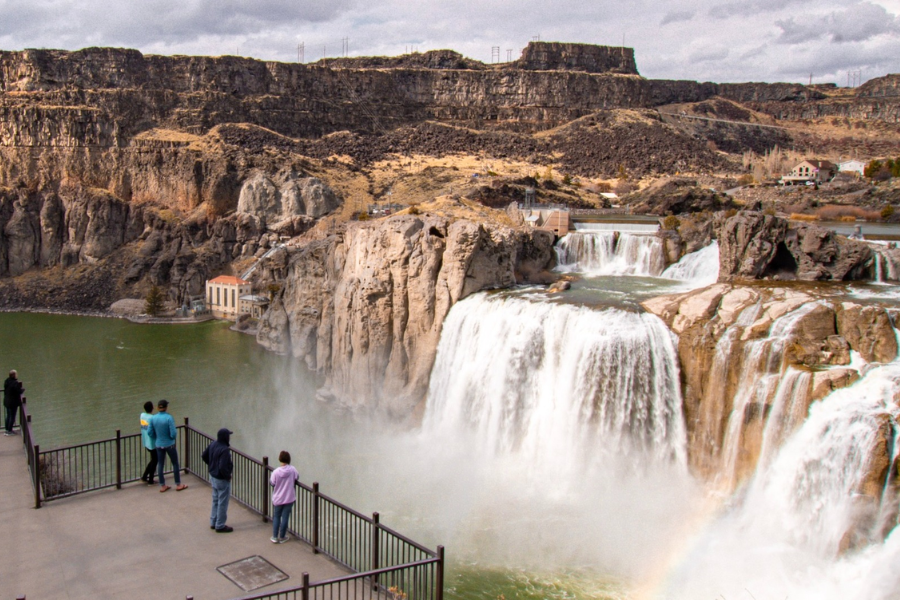Visitors view the cascading Shoshone Falls from a fenced overlook as mist rises and a faint rainbow appears near the base of the massive waterfall.