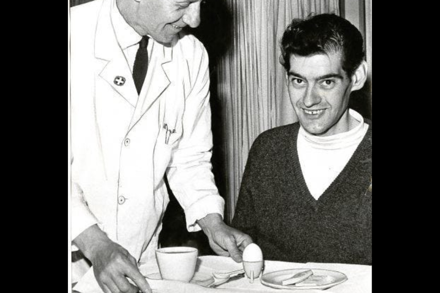 Smiling man sitting at a dining table while a doctor in a white coat serves him a light meal of tea, toast, and a soft-boiled egg.