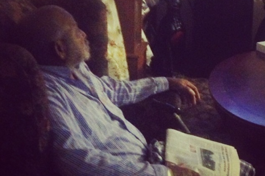 An elderly man in striped pajamas sits in a recliner, looking away while holding a folded newspaper in his lap.