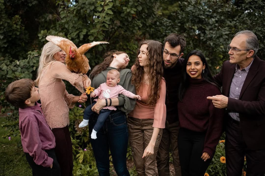 A large family gathers outdoors for a photo as a chicken flaps its wings directly in someone’s face, causing surprise and laughter all around.