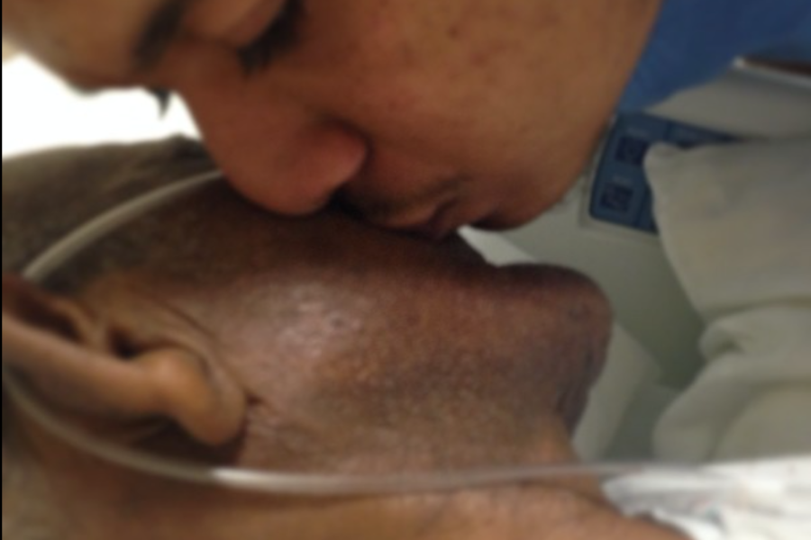 A touching close-up of a young man kissing the forehead of an elderly man in a hospital bed with an oxygen tube.