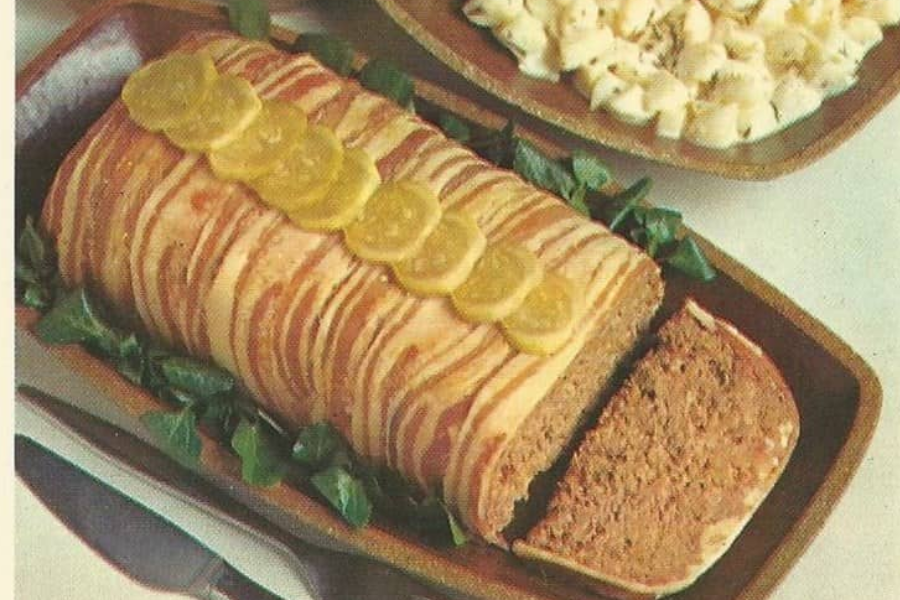 A classic meatloaf wrapped in strips of bacon, topped with pickle slices and served on a bed of greens with a side of potato salad.