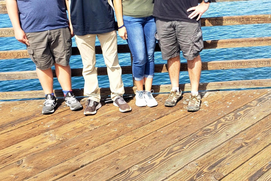 A dockside photo of four people standing side by side, but only their legs and feet are visible, cutting off everything above the waist.