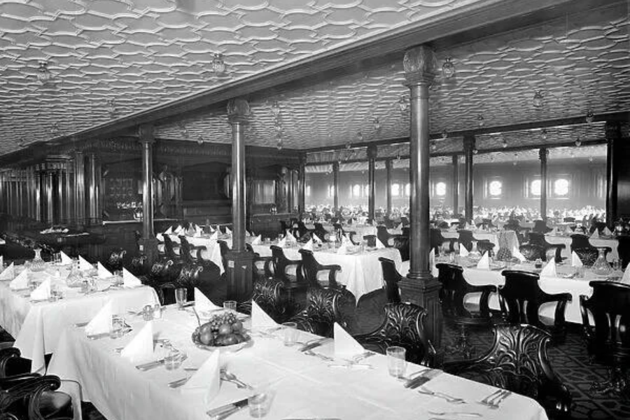 Black and white photo of Titanic's first-class dining saloon, featuring ornate wooden paneling, white tablecloths, and neatly folded napkins.