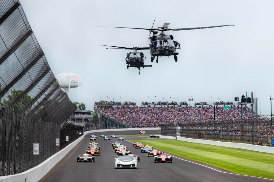 Race cars speed down the Indianapolis Motor Speedway as military helicopters fly overhead and packed grandstands cheer them on.