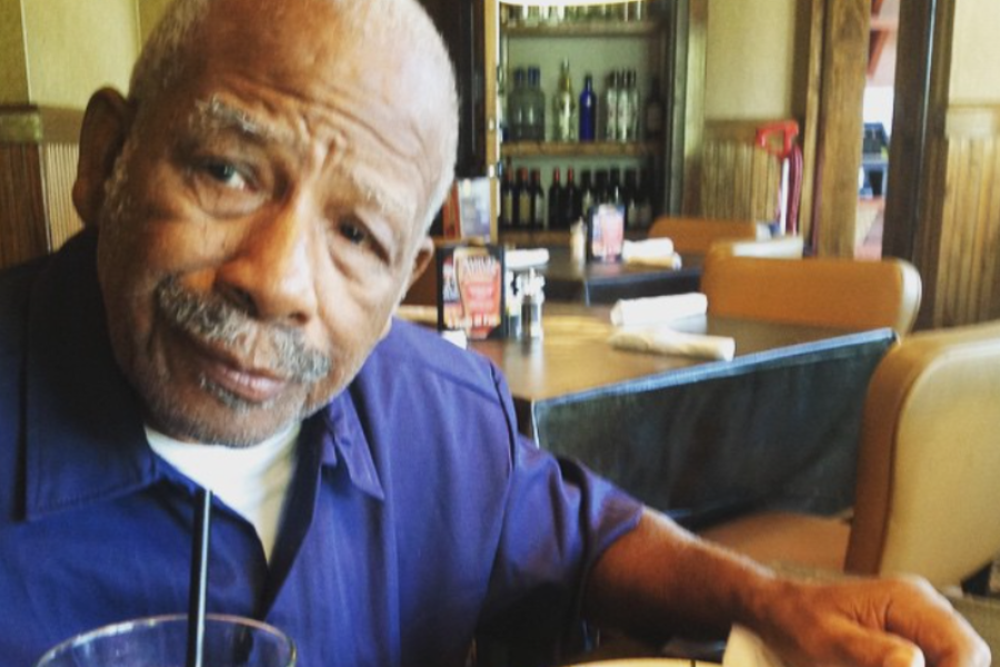 An older man in a blue shirt sits at a restaurant table, gazing thoughtfully with a drink in front of him.