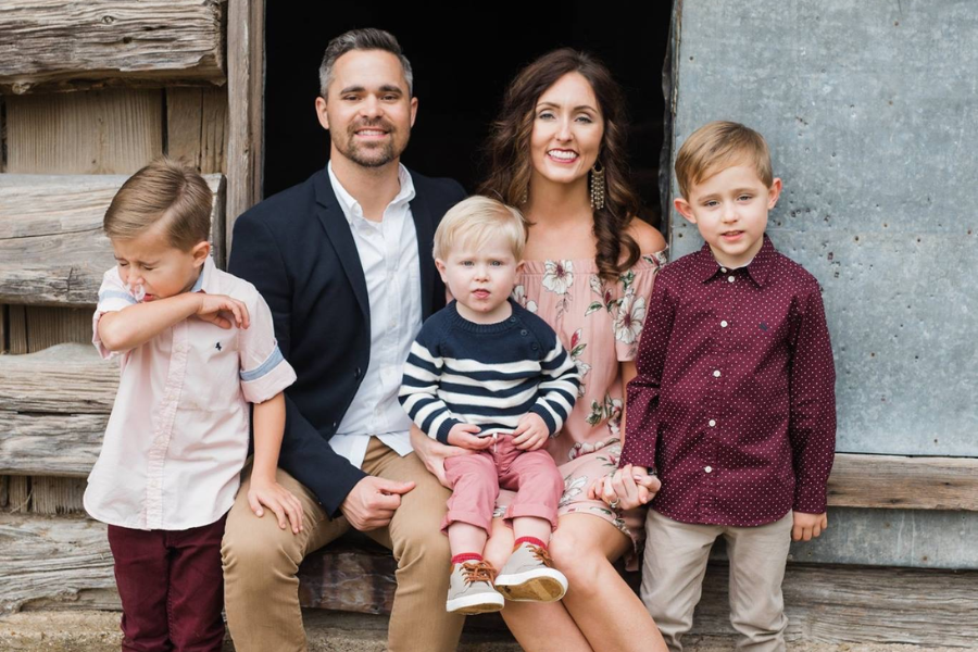 A rustic family portrait outside a barn shows everyone smiling—except one child on the left caught mid-sneeze or mid-cough.