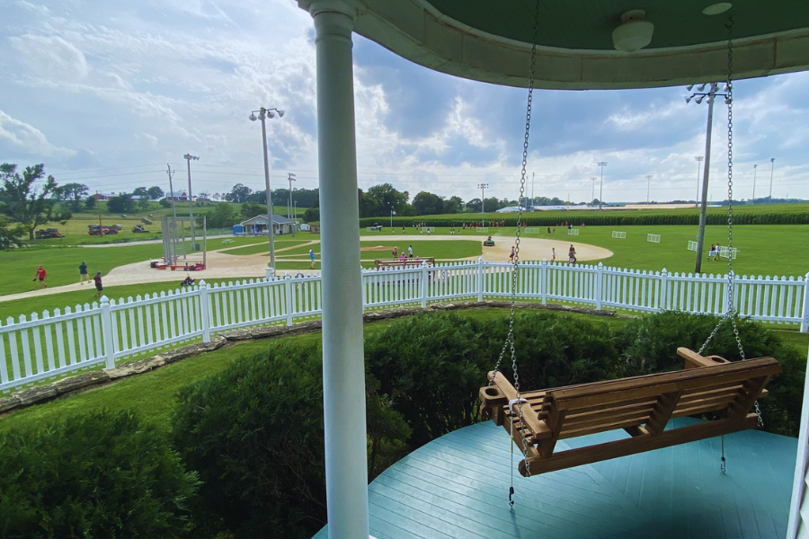 A wooden porch swing overlooks a picturesque baseball field enclosed by a white picket fence, with people playing and relaxing under a bright sky.