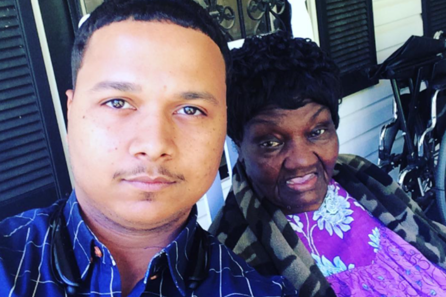 A man takes a selfie with an older woman dressed in a purple floral outfit and camouflage shawl, sitting together outside on a porch.