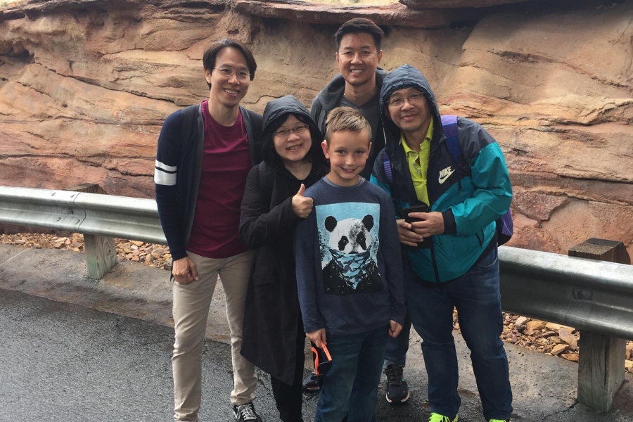 A group of five people smile for a photo on a rainy day by a guardrail and canyon wall, with one giving a thumbs-up despite the wet weather.