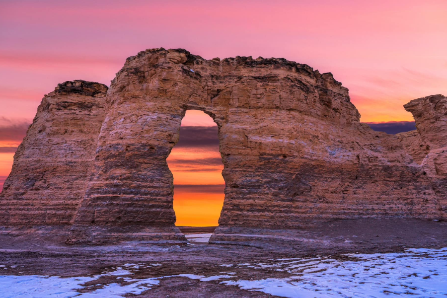 Monument Rocks, a towering natural limestone formation in Kansas, stands silhouetted against a vivid pink and orange sunset sky with patches of snow on the ground.