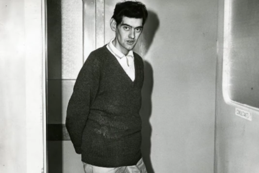 Thin man standing in a hallway with hands behind his back, wearing a dark sweater and light trousers, looking slightly serious.