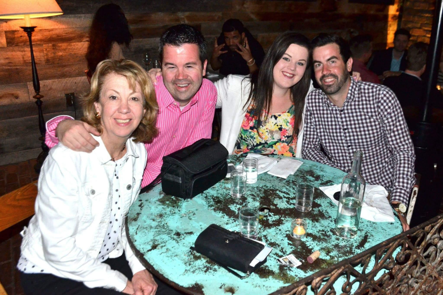 A group photo at a restaurant shows four people smiling at a table, with a man in the background flashing hand signs and photobombing.