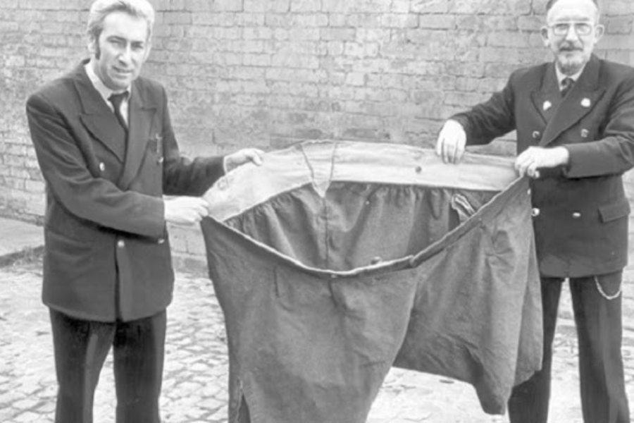 Two men holding up an enormous pair of pants outdoors, likely to illustrate the previous size of someone who experienced significant weight loss.
