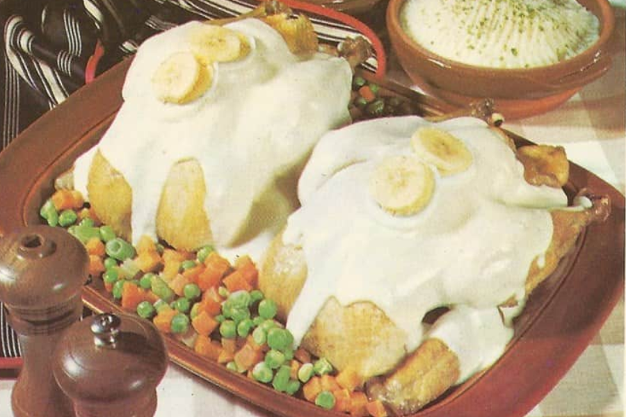 Two whole roasted chickens topped with white banana cream sauce and banana slices, served on a bed of peas and diced carrots.