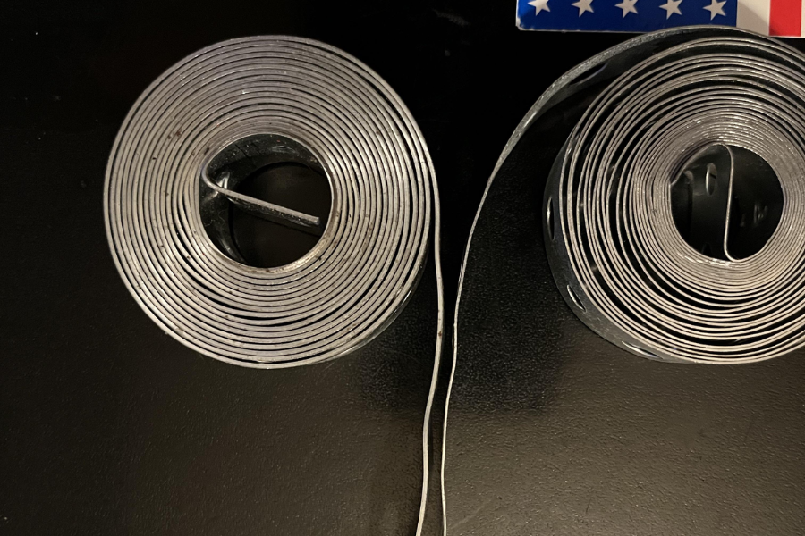 Two tightly coiled metal straps or bands placed side by side, one with a smooth edge and one with evenly spaced holes along the length.