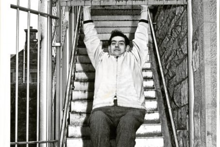 Man in a jacket hanging from a metal bar at the top of a staircase, legs lifted off the steps as part of a physical activity.