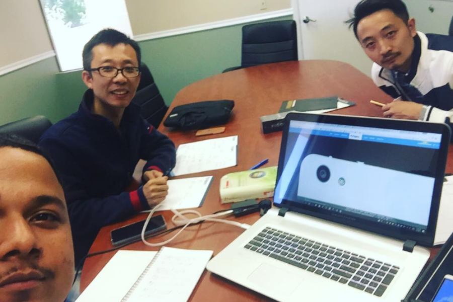 A group of three men gathered around a conference table with notebooks, electronics, and a laptop showing a tech design.