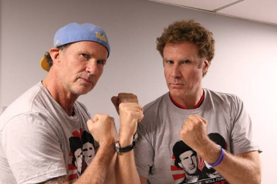Two men posing playfully with raised fists, both wearing matching gray T-shirts featuring graphic portraits and red accents. The man on the left wears a backward blue cap, while the man on the right has curly hair and a serious expression.