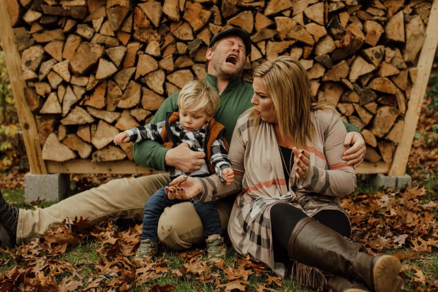 A fall-themed family photo with a woodpile backdrop shows a man wincing in pain and a woman reacting dramatically, while their toddler remains unfazed.