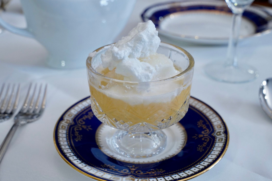 Elegant glass dessert dish of peaches in Chartreuse jelly topped with soft meringue, served on a blue-rimmed Titanic replica plate.