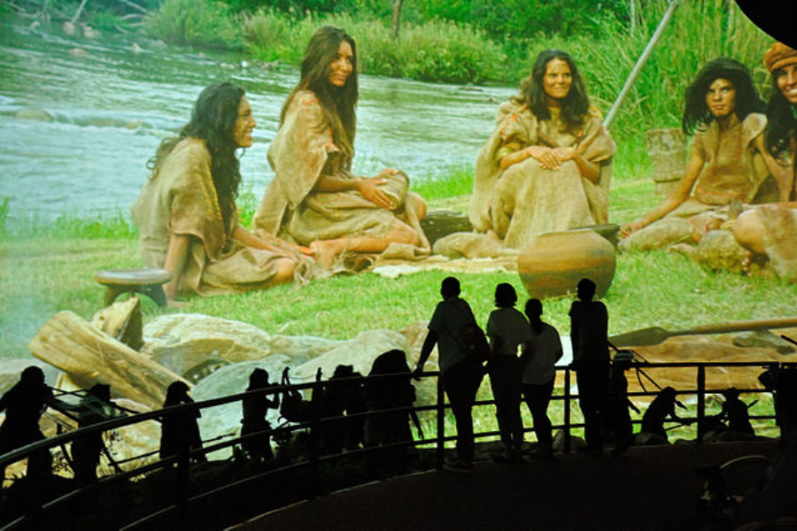 Visitors silhouetted against a large museum diorama showing prehistoric women in natural clothing gathered near a riverbank.