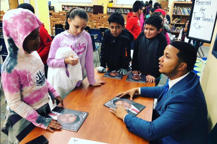 A man in a blue suit talks with a group of children holding magazines featuring his face during a school visit.