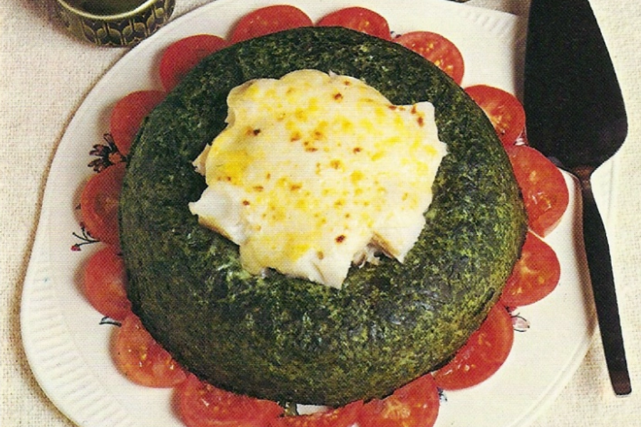A green spinach ring filled with cheesy white sauce in the center, surrounded by halved cherry tomatoes on a white plate.