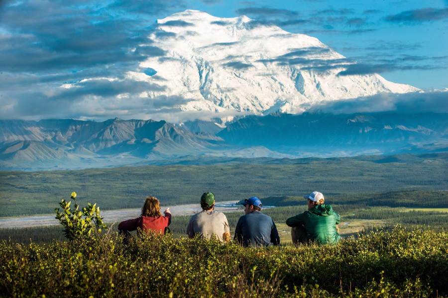 Four hikers sit on a grassy ridge overlooking a vast forested valley with Mount Denali’s snow-covered peak rising majestically in the distance.