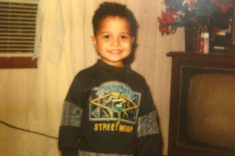 A young boy smiling brightly while wearing a “Street Wear” sweatshirt with a cartoon character design, standing indoors near a TV and air conditioner.
