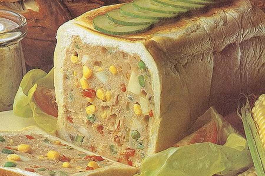 A loaf of white bread hollowed out and stuffed with a cold meat and vegetable filling, including corn, peas, and red peppers, with cucumber slices on top.