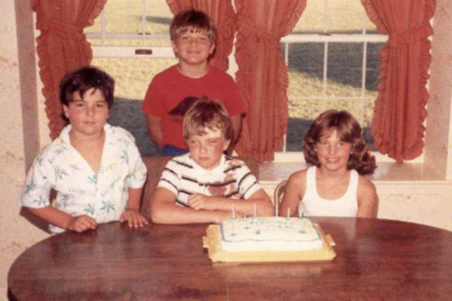 A retro birthday photo shows four kids sitting around a table with a cake, but the birthday child in the center looks visibly unhappy.