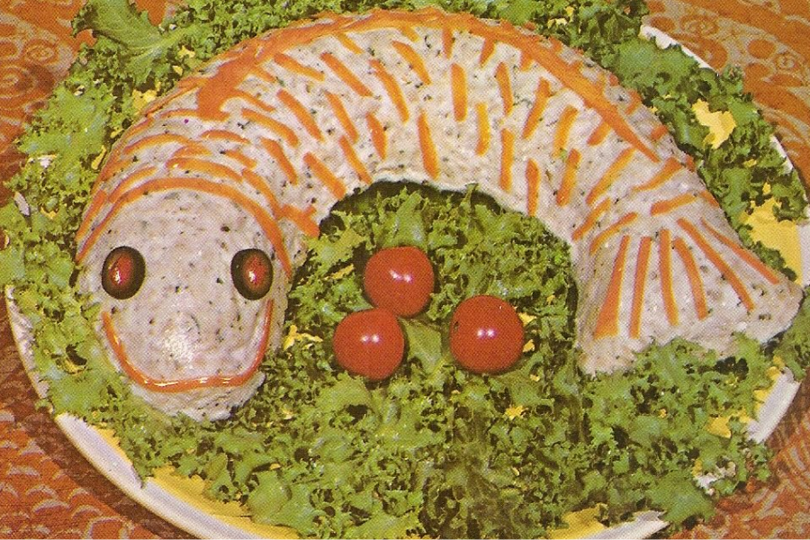 A fish-shaped molded salad decorated with olives for eyes and stripes of carrots, served on a bed of leafy greens with cherry tomatoes.