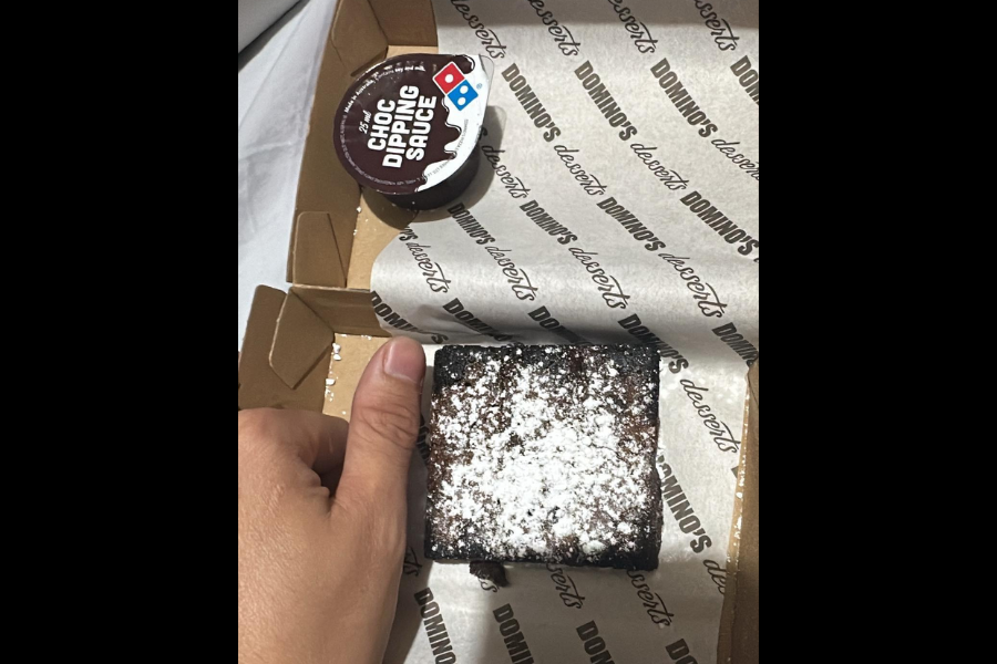 A Domino's chocolate lava cake topped with powdered sugar in a branded dessert box, with a container of Choc Dipping Sauce above it and a hand for scale.