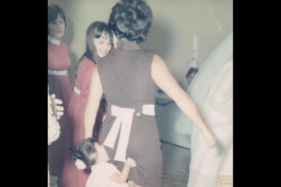 A young girl appears to be biting the back of a woman’s dress at a formal event, while others in the background react with amused smiles.