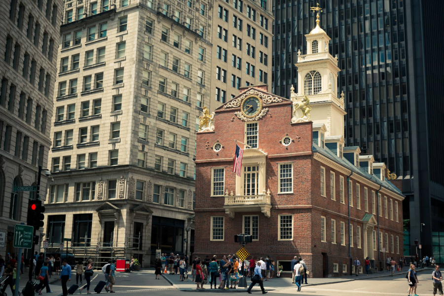 The Old State House, a red brick colonial-era building with a white steeple and gold accents, stands prominently among modern skyscrapers in downtown Boston.