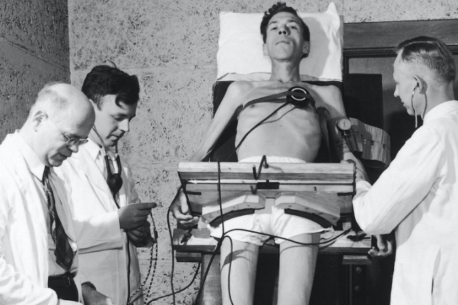 Black and white photo of a thin man strapped to a medical tilt table with three doctors examining him, likely during a fasting or cardiovascular study.