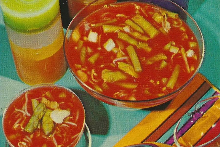 Bowls of bright red tomato-based soup filled with green beans, sprouts, and chopped vegetables, served alongside a multicolored layered gelatin drink.