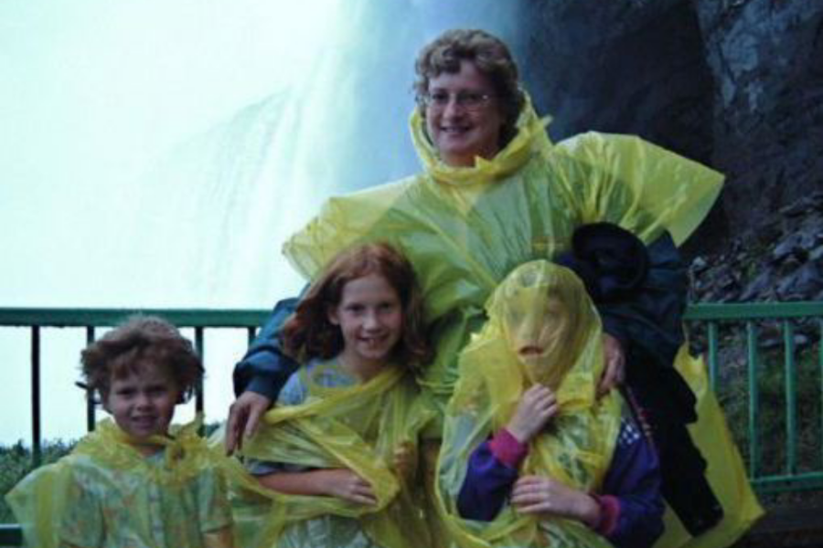 A woman and three children wear oversized yellow ponchos in front of a waterfall; one child has the hood covering their entire face.