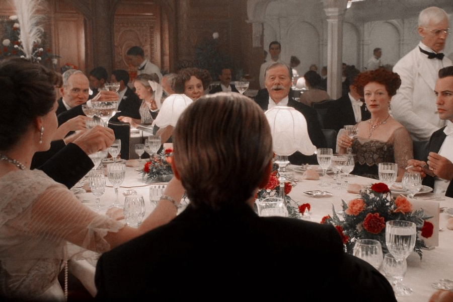 Formal dinner scene from the movie Titanic with well-dressed passengers seated around an elegant first-class table adorned with crystal glassware and floral arrangements.