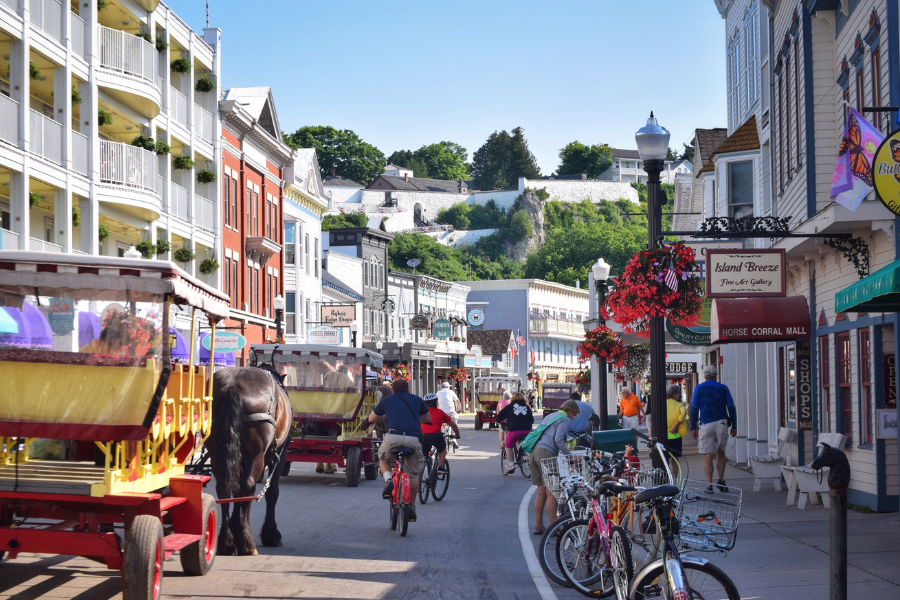 A lively street scene on Mackinac Island features bicycles, horse-drawn carriages, and charming historic buildings, with Fort Mackinac perched on a hill in the background.