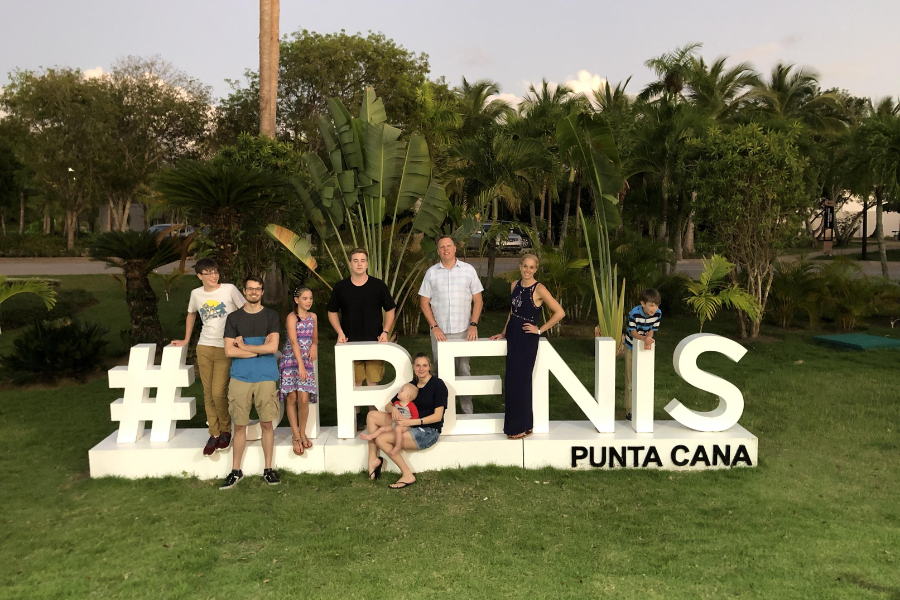 A group poses in front of a large white "#PUNTA CANA" sign, but their placement blocks letters, humorously leaving the word “#PENIS.”