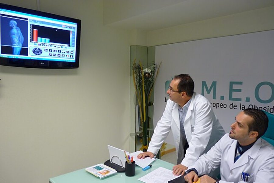 Two male doctors in lab coats examine a digital scan of a human body on a wall-mounted screen inside a clinic with “C.M.E.O.” signage.