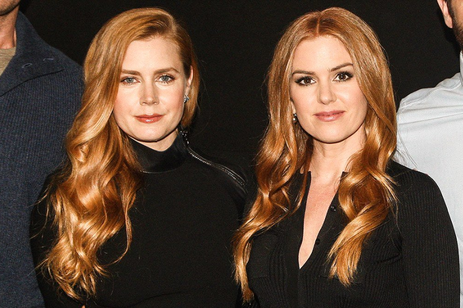 Side-by-side photo of two women with long, wavy red hair, both wearing black outfits and posing with neutral expressions. They are standing close together against a dark background.